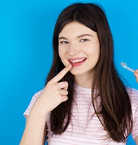Teen with dark hair pointing to smile with one hand holding Invisalign in the other